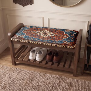 May include: A wooden entryway bench featuring a rectangular seat cushion with a blue, orange, and cream-colored ornate pattern. The bench has a slatted lower shelf holding a pair of white sneakers and a pair of brown leather shoes.