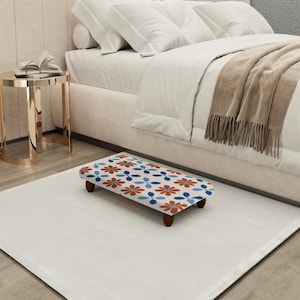 May include: A floral patterned footstool with a white background, featuring a repeating design of orange and blue flowers. The footstool has four wooden legs and is placed on a white rug near a bed.