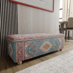 Op de afbeelding: Een rechthoekige opbergottoman met een gedetailleerd, kleurrijk patroon. Het ontwerp heeft een turquoise, roze en oranje bloemenmotief. De ottoman staat op korte, lichtgekleurde houten poten en staat op een houten vloer.