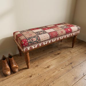 Puede incluir: Un banco rectangular con un asiento acolchado tapizado en una tela de retazos multicolor con patrones florales en rojo, verde y crema. El banco se apoya sobre cuatro patas de madera torneada con un acabado en marrón cálido.