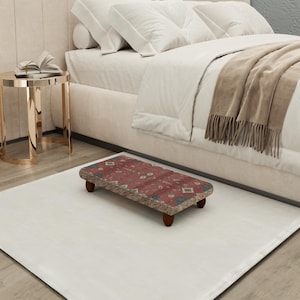 May include: A small, rectangular footstool with a patterned rug top in shades of red, brown, and green. The footstool has four short, dark wooden legs and is placed on a white rug near a bed.