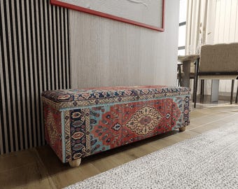 Puede incluir: Un banco de almacenamiento rectangular tapizado con una tela que presenta un patrón intrincado multicolor en tonos rojo, azul y crema. El banco tiene una parte superior plana, lados rectos y se apoya sobre cuatro pequeñas patas cilíndricas de madera clara.