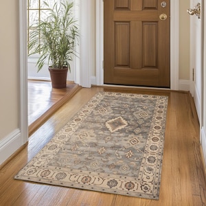 Puede incluir: Una alfombra rectangular estampada en un pasillo. La alfombra presenta un diseño central en tonos grises y beige, con un borde de motivos florales repetidos. La alfombra está colocada frente a una puerta de madera y sobre un suelo de madera.