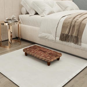 Puede incluir: Un pequeño reposapiés rectangular con una parte superior de alfombra estampada en tonos rojos, marrones y verdes. El reposapiés tiene cuatro patas de madera oscura y está colocado sobre una alfombra blanca cerca de una cama.