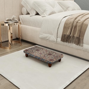 May include: A rectangular footstool with a patterned rug design, featuring a gray background with red and blue floral accents. The footstool has dark wooden legs and is placed on a white rug near a bed.