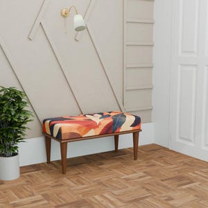 May include: A decorative bench with a patterned seat in shades of orange, red, and blue. The bench has a wooden frame and legs, and is set against a neutral wall. A potted plant and a wall light are also visible. The bench is approximately 100 cm long.