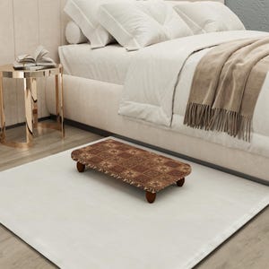 May include: A rectangular, brown footstool with a patterned design, resting on four short, dark wooden legs. The footstool is placed on a white rug in front of a bed with white bedding and a tan throw blanket.