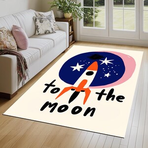 以下が含まれることがあります： ロケットと星が描かれた青とピンクの円が描かれた白い敷物。円の下に黒い文字で「to the moon」と書かれています。
