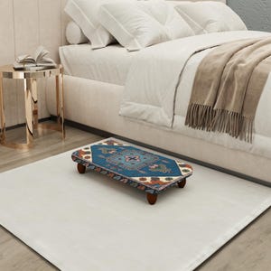May include: A small, rectangular footstool with a patterned top in shades of blue, red, and beige. The footstool has four short, dark wooden legs. It is placed on a white rug near a bed with white bedding.