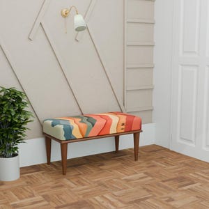May include: A colourful patterned bench with wooden legs, set against a neutral wall and parquet flooring. The seat features an abstract design in shades of orange, blue, and yellow. A potted plant and a wall light are also in the room.