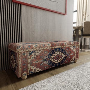 May include: A rectangular storage ottoman with a patterned fabric in shades of red, blue, and cream. The ottoman has a wooden frame and short, rounded wooden legs. The design features a traditional rug-like pattern.
