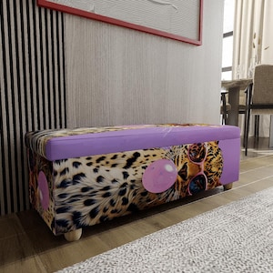 May include: A rectangular storage ottoman featuring a leopard print pattern with a graphic of a leopard wearing pink sunglasses and blowing a pink bubblegum bubble. The top lid is solid purple, and the base rests on four small light wooden feet.