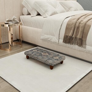 May include: A patterned footstool with dark wooden legs rests on a white rug. The footstool has a rectangular shape with rounded edges and a muted colour palette of blues, greys, and beige. The footstool is near a bed.