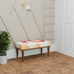 Pode incluir: Banco decorativo com uma almofada creme com formas abstratas laranja e amarelas. O banco tem uma estrutura e pernas de madeira. Um vaso de plantas e uma luminária de parede também são visíveis.