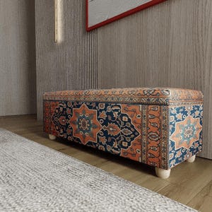 Op de afbeelding: Een rechthoekige poef met een patroonstof in tinten blauw, oranje en crème. De poef heeft een houten frame en korte, lichtgekleurde poten. Het ontwerp bevat geometrische en bloemmotieven.