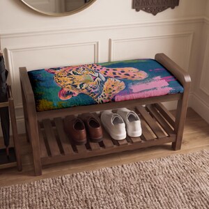 May include: A wooden entryway bench with a rectangular upholstered seat featuring a colorful leopard print in shades of blue, pink, and yellow. The bench includes a lower slatted shelf for storage, currently holding a pair of brown shoes and white sneakers.