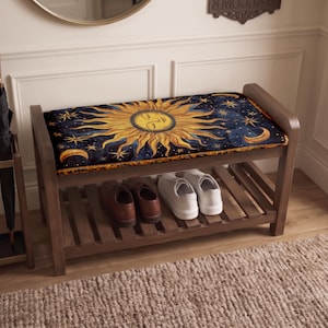 May include: A wooden entryway bench featuring a cushioned seat with a celestial print of a golden sun, crescent moon, and stars against a dark blue background. The bench has a slatted lower shelf holding two pairs of shoes, one brown and one white.