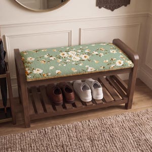 Può includere: Una panca scarpiera in legno con un cuscino rettangolare in tessuto verde salvia con un motivo floreale bianco e giallo. La panca presenta un ripiano inferiore a doghe che ospita un paio di scarpe in pelle marrone e un paio di scarpe da ginnastica.