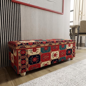 Könnte beinhalten: Ein rechteckiger Aufbewahrungs-Hocker mit einer gemusterten Stoffoberfläche. Das Design zeigt rote, blaue und cremefarbene Quadrate mit floralen Motiven. Der Hocker hat Holzfüße und steht in einem Raum mit neutralem Farbschema.