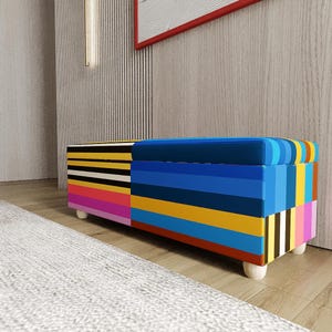 May include: A multi-coloured storage bench with a striped pattern. The bench incorporates blue, yellow, pink, and black. It has a rectangular form and is supported by four small, rounded wooden legs. The bench is positioned on a wooden floor.