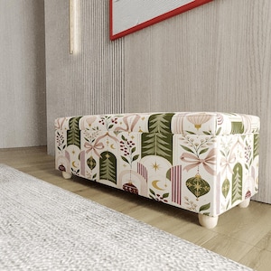 Puede incluir: Un banco de almacenamiento rectangular tapizado en tela con un patrón repetitivo de pinos verdes, lazos rosas, adornos dorados y bayas sobre un fondo crema, sostenido por cuatro pequeñas patas de madera de color claro.