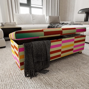 May include: A colorful storage ottoman with a striped pattern in red, yellow, pink, and green. The lid is open, revealing a dark gray blanket inside. The ottoman sits on a light-colored rug in a modern living room setting.