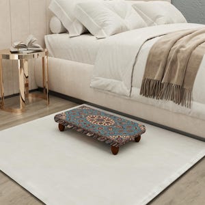 Puede incluir: Un pequeño reposapiés rectangular con una parte superior estampada en tonos azules, marrones y crema. El reposapiés tiene cuatro patas cortas de madera oscura y está colocado sobre una alfombra blanca cerca de una cama.