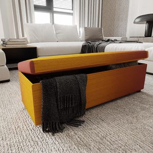 May include: A rectangular storage bench with a hinged lid, featuring a warm yellow and orange color scheme. The bench is open, revealing a dark gray blanket inside. The lid has a pink edge.