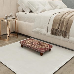 Puede incluir: Un reposapiés rectangular con un diseño de alfombra estampada en tonos rojos, beige y marrón. El reposapiés tiene cuatro patas cortas de madera oscura y está colocado sobre una alfombra blanca cerca de una cama.