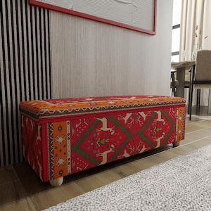 May include: A rectangular storage ottoman with a vibrant red, orange, and green patterned fabric. The design features geometric shapes and animal motifs. The ottoman rests on four short, light-coloured wooden legs, adding a touch of elegance to the piece.