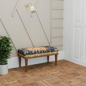 May include: A decorative bench with a celestial design. The bench features a dark blue cushion with a gold sun and moon pattern. It has four wooden legs and is placed against a neutral-colored wall. A potted plant and a wall sconce are also visible.