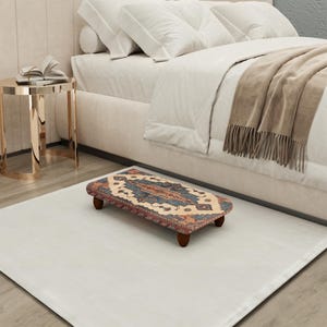 May include: A small, rectangular footstool with a patterned rug top in shades of blue, red, and beige. The footstool has four short, dark wooden legs. It is placed on a white rug near a bed with white bedding.