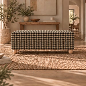 May include: A rectangular storage ottoman upholstered in a black and white houndstooth fabric pattern. The piece features a flat top surface and is supported by four rounded, light-colored wooden feet positioned at each corner of the base.