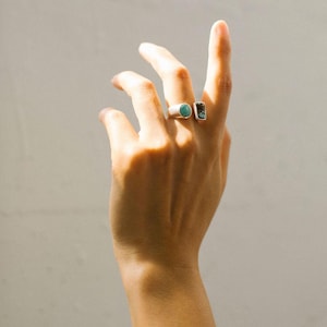 May include: A hand wearing two silver rings with turquoise stones. The ring on the index finger has a round stone, and the ring on the middle finger has a rectangular stone.