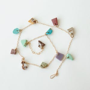 Puede incluir: Un collar de cadena fina en tono dorado con m&uacute;ltiples dijes peque&ntilde;os de formas irregulares en varios colores, incluyendo turquesa, lavanda, terracota, marr&oacute;n y verde, distribuidos uniformemente a lo largo de la cadena de eslabones.