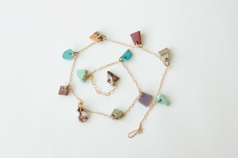 Puede incluir: Un collar de cadena fina en tono dorado con m&uacute;ltiples dijes peque&ntilde;os de formas irregulares en varios colores, incluyendo turquesa, lavanda, terracota, marr&oacute;n y verde, distribuidos uniformemente a lo largo de la cadena de eslabones.