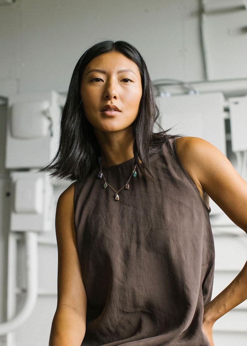 Puede incluir: Una blusa sin mangas de color marr&oacute;n oscuro con cuello alto combinada con un delicado collar de cadena dorada que presenta peque&ntilde;os colgantes de piedras preciosas de formas irregulares en tonos morado, verde y rosa.