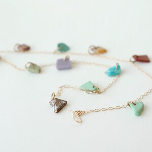 Puede incluir: Un collar de cadena fina en tono dorado con m&uacute;ltiples dijes de piedra de formas irregulares en varios colores, incluyendo turquesa, lavanda, verde menta, marr&oacute;n y blanco, distribuidos uniformemente a lo largo de la cadena de eslabones.