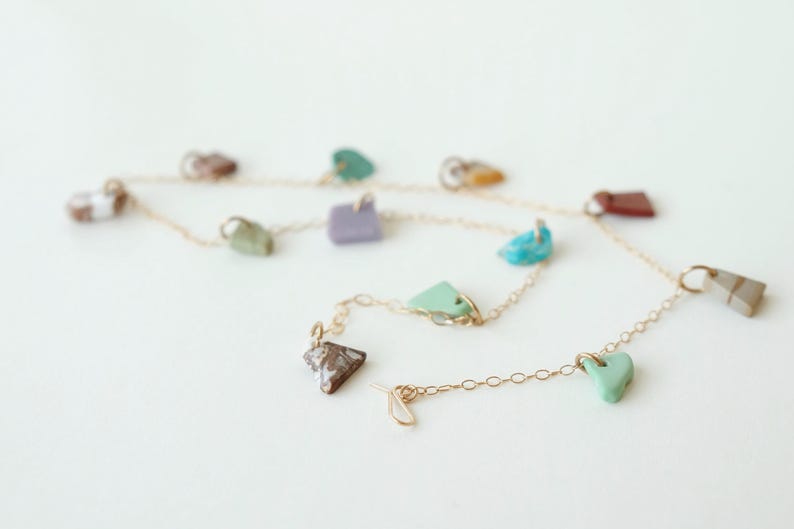 Puede incluir: Un collar de cadena fina en tono dorado con m&uacute;ltiples dijes de piedra de formas irregulares en varios colores, incluyendo turquesa, lavanda, verde menta, marr&oacute;n y blanco, distribuidos uniformemente a lo largo de la cadena de eslabones.