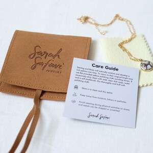 Puede incluir: Un collar de cadena dorada con un colgante redondo de patr&oacute;n marr&oacute;n y blanco, acompa&ntilde;ado de una bolsa de joyer&iacute;a de ante color canela con el sello Sarah Safari Jewelry, un pa&ntilde;o de pulido amarillo p&aacute;lido y una tarjeta de cuidados impresa.