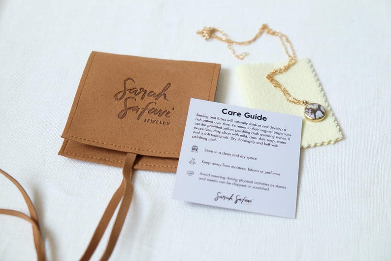 Puede incluir: Un collar de cadena dorada con un colgante redondo de patr&oacute;n marr&oacute;n y blanco, acompa&ntilde;ado de una bolsa de joyer&iacute;a de ante color canela con el sello Sarah Safari Jewelry, un pa&ntilde;o de pulido amarillo p&aacute;lido y una tarjeta de cuidados impresa.