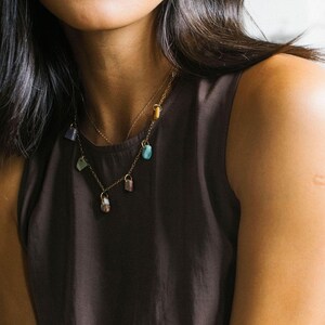 Puede incluir: Un collar de cadena en tono dorado con varias peque&ntilde;as cuentas de piedras preciosas de formas irregulares en tonos azul, morado, verde y marr&oacute;n, suspendidas de delicados eslabones met&aacute;licos sobre una blusa sin mangas de color marr&oacute;n oscuro.