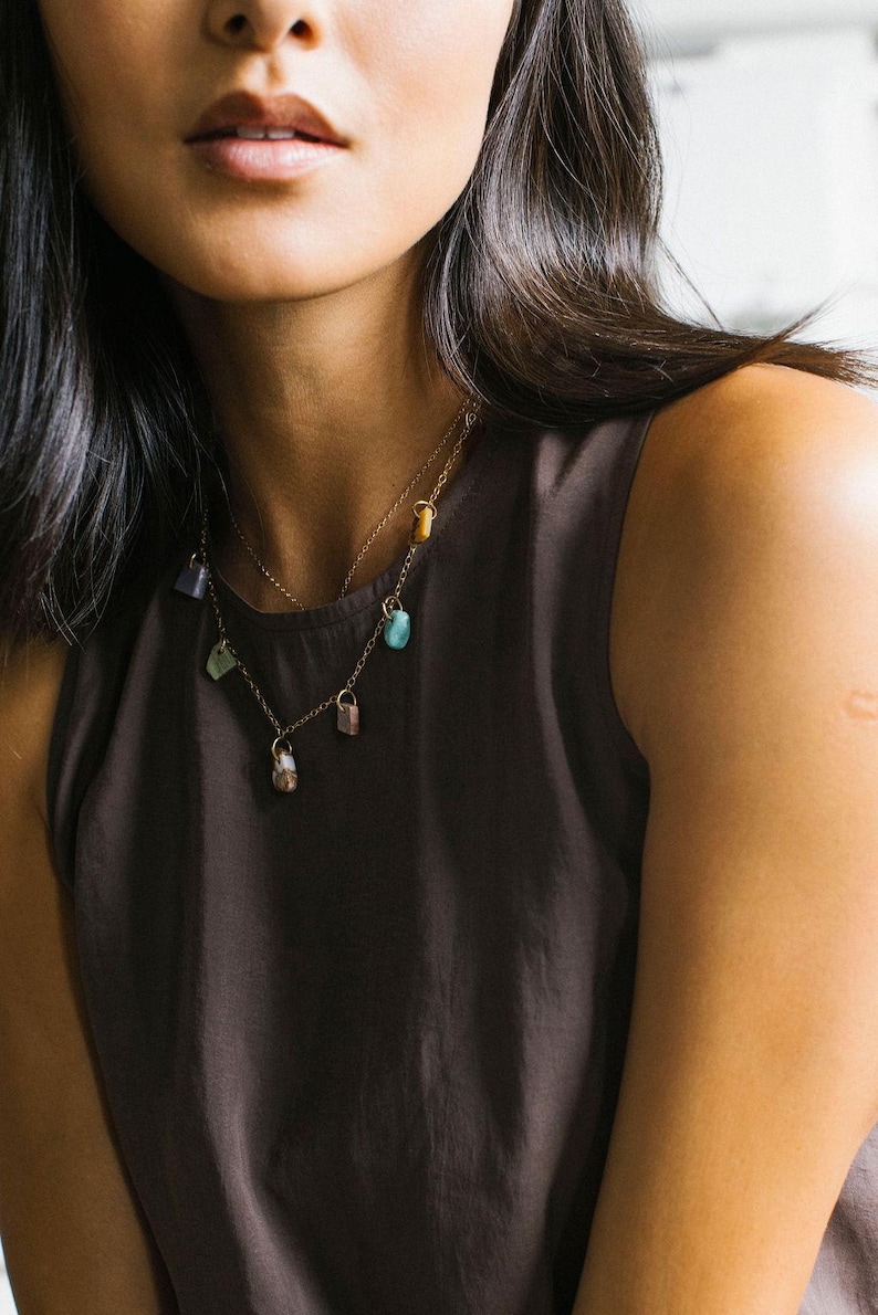 Puede incluir: Un collar de cadena en tono dorado con varias peque&ntilde;as cuentas de piedras preciosas de formas irregulares en tonos azul, morado, verde y marr&oacute;n, suspendidas de delicados eslabones met&aacute;licos sobre una blusa sin mangas de color marr&oacute;n oscuro.