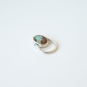 Puede incluir: Un anillo de plata con una piedra turquesa de forma ovalada con inclusiones de matriz marrón. La piedra está engastada en un bisel de metal liso y la banda tiene un diseño redondeado y abierto que se curva alrededor del engaste en un bucle.