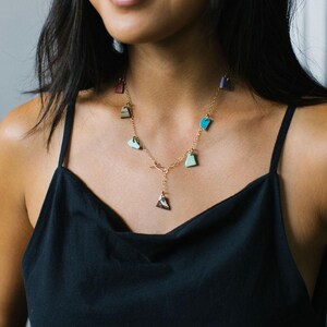 Puede incluir: Un collar de cadena dorada con m&uacute;ltiples dijes peque&ntilde;os de formas irregulares en varios colores, incluyendo turquesa, verde claro, rojo oscuro y morado, con un colgante central triangular de patr&oacute;n moteado marr&oacute;n y blanco.