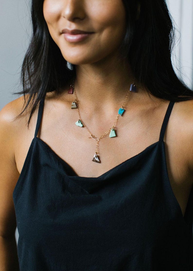 Puede incluir: Un collar de cadena dorada con m&uacute;ltiples dijes peque&ntilde;os de formas irregulares en varios colores, incluyendo turquesa, verde claro, rojo oscuro y morado, con un colgante central triangular de patr&oacute;n moteado marr&oacute;n y blanco.