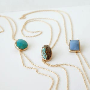 Puede incluir: Tres collares dorados con colgantes distintos: una piedra turquesa redonda, una piedra marrón ovalada con motas turquesas y una piedra azul claro rectangular. Cada colgante está engastado en un marco de metal dorado sobre una cadena fina.
