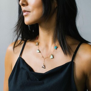 Puede incluir: Un collar de cadena en tono dorado con m&uacute;ltiples dijes peque&ntilde;os de formas irregulares en varios colores, incluyendo turquesa, verde claro y marr&oacute;n oscuro. El collar tiene un dise&ntilde;o de eslabones delicados con un dije central y cierre de barra.