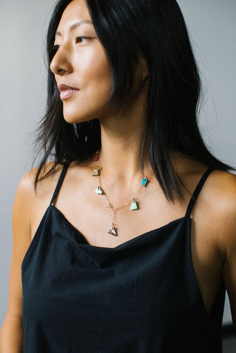 Puede incluir: Un collar de cadena en tono dorado con m&uacute;ltiples dijes peque&ntilde;os de formas irregulares en varios colores, incluyendo turquesa, verde claro y marr&oacute;n oscuro. El collar tiene un dise&ntilde;o de eslabones delicados con un dije central y cierre de barra.