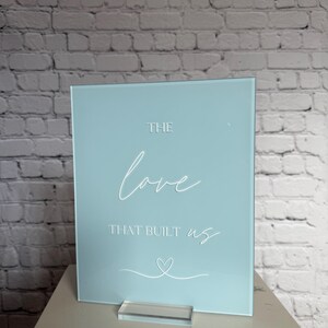 May include: A light blue acrylic sign with the words "THE love THAT BUILT us" in white script. A white heart is drawn below the text. The sign is displayed on a clear acrylic stand.